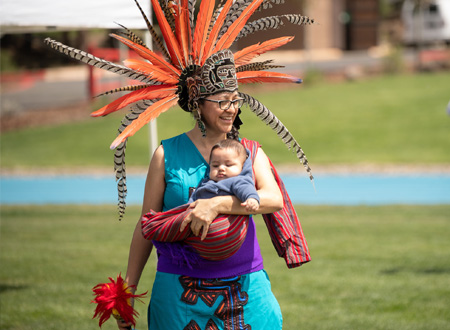 Smiling Female Native American Dancer in Regalia Holding Baby in Fabric Sling