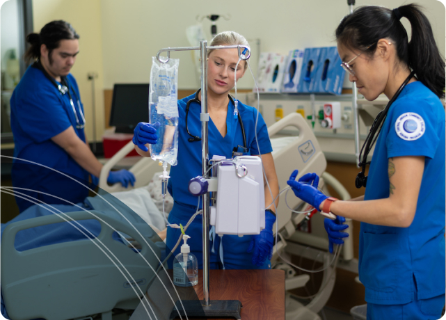 Three nursing students in blue scrubs working in a hands-on-learning setting, with one adjusting an IV setup while the others assist.