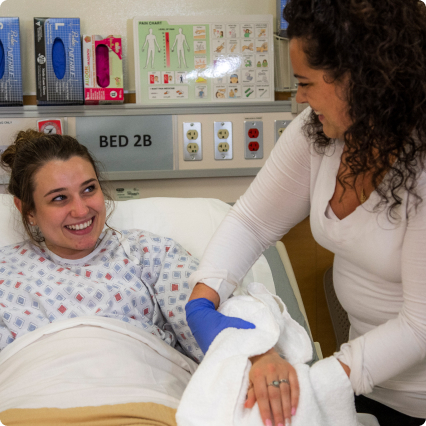 A nursing student in grey scrubs assists a student patient actor in teal scrubs by pricking their finger for a blood sample, both smiling during the interaction.