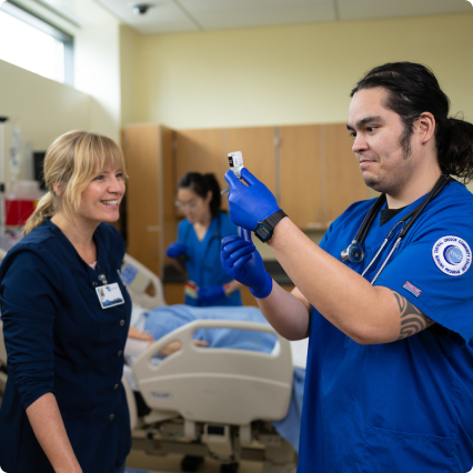 A nursing student in blue scrubs holds a medical vial while showing it to an instructor in a navy uniform, with a patient bed and another student visible in the background.