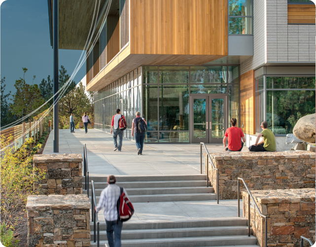 Students walk towards the entrance of the Middleton Science Center at Central Oregon Community College (COCC). The building features modern wooden accents and large glass windows. Two students are seated on stone benches near the entrance, and the pathway is lined with trees, offering a scenic view. The day is bright and sunny, adding to the welcoming campus atmosphere.