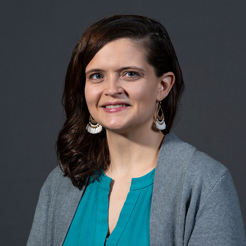 Headshot of Heather smiling directly at the camera wearing a teal blouse and gray cardigan on a dark gray backdrop.